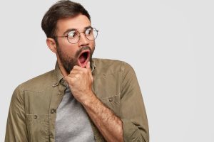 Scared anxious male keeps hand near opened mouth, looks with very frightened expression aside, notices something awful, wears round spectacles and fashionable shirt, poses in studio against white wall